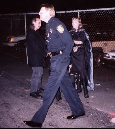 Black-clad policeman strides by black-clad Witches at Coven Oldenwilde's Samhain 97 free public Witch ritual at Asheville's Memorial Stadium