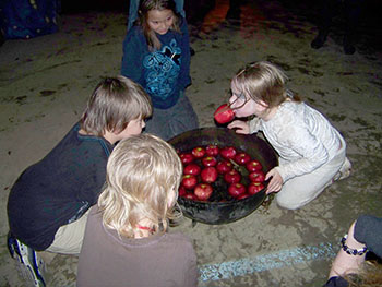 Kids surrounding cauldron full of floating apples, one with apple in her mouth.