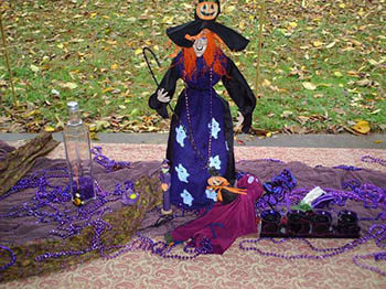 Witch figurine on purple altar against fallen leaves on grass.