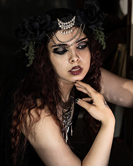 A young dark-haired female Witch wearing a crescent-moon tiara and a thoughtful expression holds her fingertips below her chin